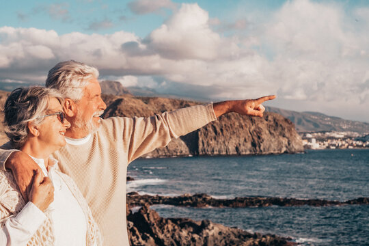 Senior couple embracing and smiling on a rocky coastline, looking at the ocean and distant town while the man points towards the horizon, symbolizing a shared future and adventure together