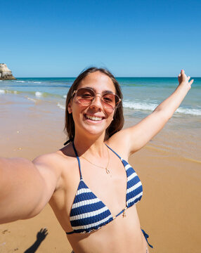 A young woman smiles and raises her arms while taking a selfie on a sunny sandy beach, wearing sunglasses and a blue striped bikini with gentle ocean waves and clear sky behind her.