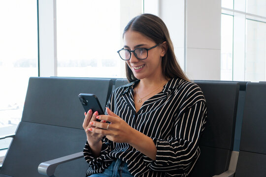 Young woman traveler wearing glasses and a striped shirt, sitting on an airport terminal bench, smiling and using her mobile phone while waiting for her flight
