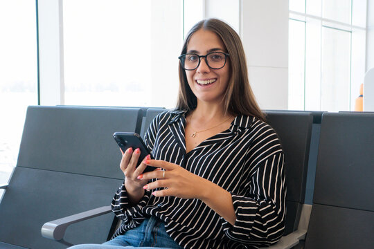 A smiling young woman in glasses sits in an airport gate holding her smartphone, dressed casually and appearing relaxed and ready for travel, remote work or checking messages before boarding.