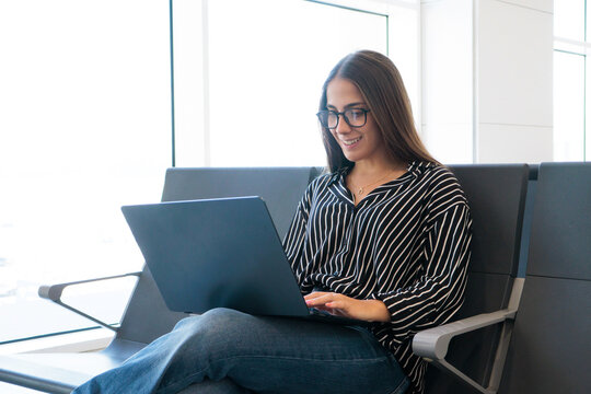 Young woman with glasses smiles as she works on a laptop in an airport terminal, combining travel and remote work while waiting for a flight in a bright, modern seating area.