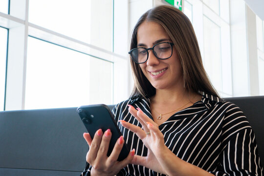 Young woman wearing glasses and striped shirt happily browsing her mobile phone, connecting with others or managing travel plans while waiting in a bright, contemporary airport terminal