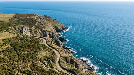 Fototapeta premium Aerial view of dramatic coastal cliffs, scenic dirt road winding along ocean coastline, blue sea water backdrop for travel advertisement