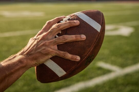 Close-Up of an Athlete's Hand Gripping an American Football Against a Sunny Outdoor Field, Detailed Sports Equipment and Ball Texture, Competitive Game, Training and Athletic Performance Concept