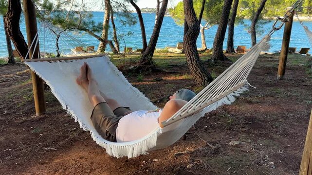 Side view of a man lying down and relaxing in a white mesh hammock under pine tree shade on a sunny coast near blue sea water.