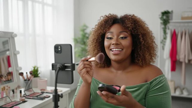 curvy black woman applying face powder with cosmetic brush while streaming live makeup tutorial on smartphone
