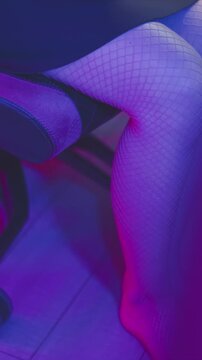 Woman legs in fishnet stockings sit on black and pink grid patterned office chair with rolling wheels illuminated by vibrant purple blue lighting. scene focuses on lower body and furniture
