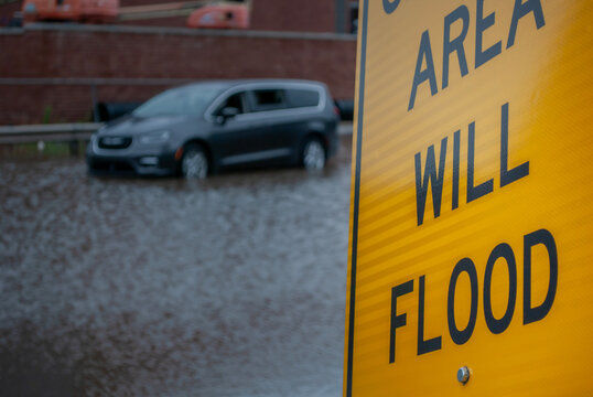 A Yellow Flood Warning Sign in Shallow Focus with a Stranded Gray Minivan Submerged on a Flooded Urban Street