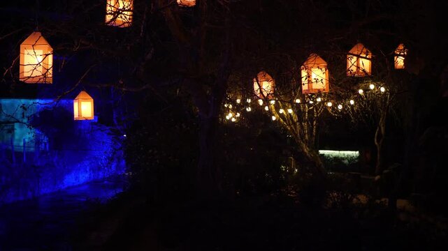 Nighttime riverside scene with glowing halloween lanterns suspended in bare trees and warm white string lights reflected in still water. Blue uplighting illuminates stone structure on left.