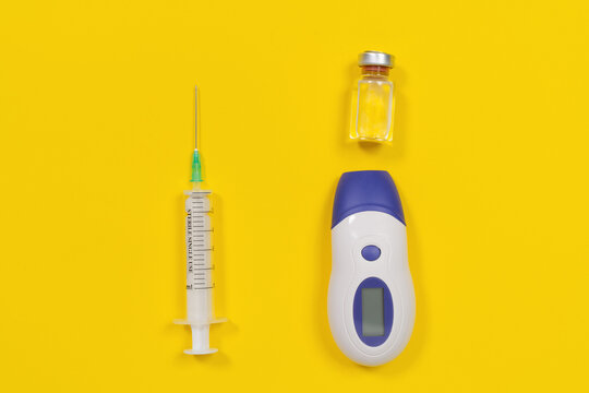 A medical syringe, a glass vaccine vial and a digital infrared thermometer are arranged flat on a bright yellow background, a concept of immunization and health monitoring.