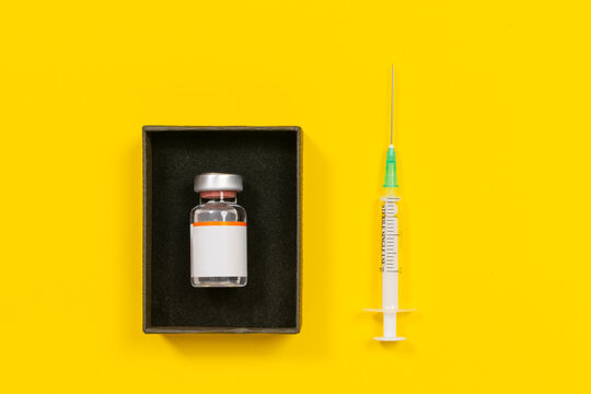 A sterile single use syringe with a green hub needle lies next to a glass medication vial with a blank orange striped label seated in a black gift style box on a bright yellow surface, a concept of.