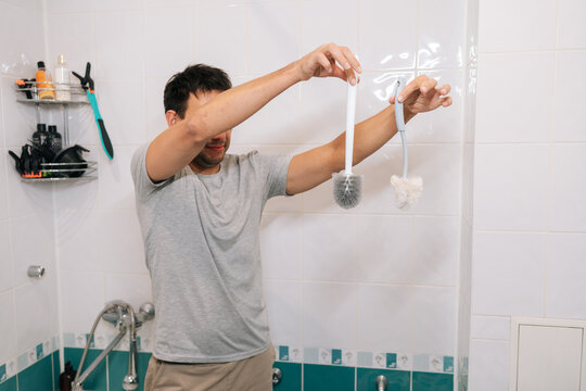 Young adult man comparing new and worn toilet brushes in bathroom, evaluating hygiene and cleanliness for routine household maintenance and toilet cleaning tools during daytime indoor life