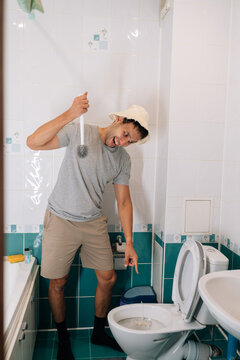 Vertical shot of cheerful young man in bucket hat holding toilet brush, making funny face pointing at dirty toilet, showing humorous take on housework chores and hygiene