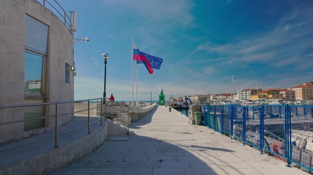 POV walking shot along the pier in Piran marina, Slovenia, showcasing scenic harbor views with flags and lighthouses under a bright blue sky.