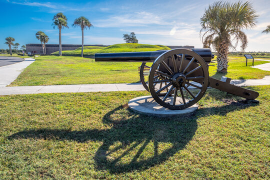 A historic cannon overlooks the grassy earthworks and brick walls of Fort Pulaski, with palm trees framing the coastal fortification.