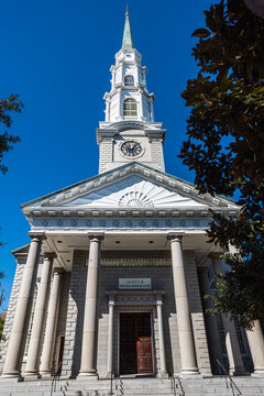 White historic church facade with classical columns, a clock tower, and a tall steeple beneath a clear blue sky.
