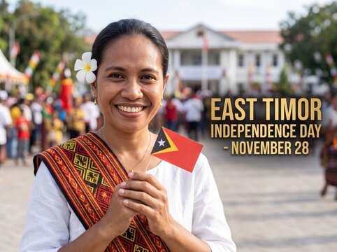 Smiling woman wearing traditional tais fabric sash holding a small timor leste flag outdoors with a blurred building and crowd in the background during a national celebration