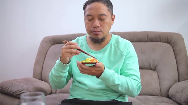 Young man Enjoy eating fresh salad with chopsticks and drinking water on cozy couch at home