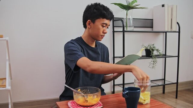 Asian Teenager reading book while snacking on crisp crackers at home