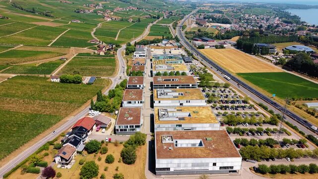 Business rooftops and parking lots border vineyard fields near Rolle, drone rising skyward