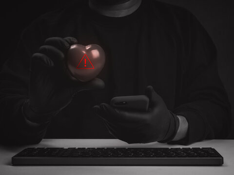 Anonymous scammer holds black heart with red warning symbol while uses smartphone. Medical Identity theft, romance scams, online dating fraud, catfishing, emotional manipulation and digital deception.