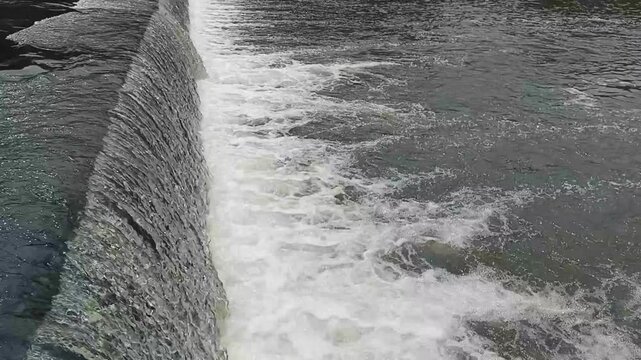 Top view of freshwater flows over a concrete weir barrier, creating foamy white rapids and steady current movement. Useful for hydrology, water management, and environmental concepts