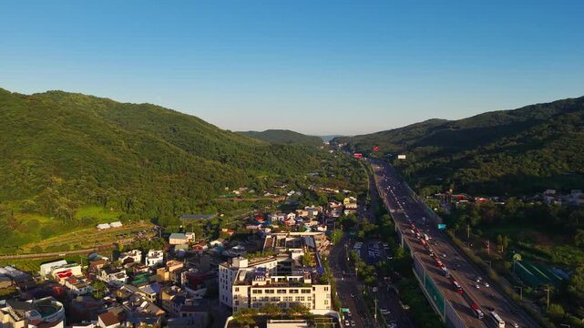 Aerial view flying forward above Gyeongbu Expressway in Seocho gu, Seoul, South Korea, with buses and cars heading into a green valley past roadside village and garden plots