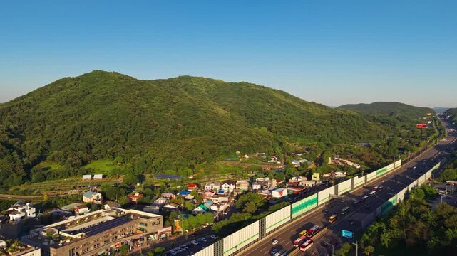 Aerial pull back reveals busy traffic Gyeongbu Expressway in Seocho gu district in Seoul with forested Cheonggyesan rising above a roadside village, garden plots