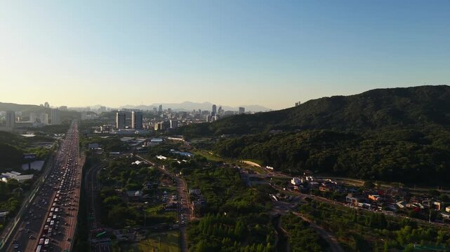 Aerial view flying over Gyeongbu Expressway in Seocho gu, Seoul, South Korea, with heavy traffic, wooded Umyeonsan slopes and hazy downtown skyline at golden hour