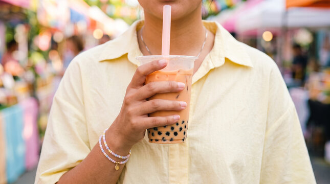 Young woman holding orange boba milk tea with black tapioca pearls in clear plastic cup at vibrant outdoor street food market.