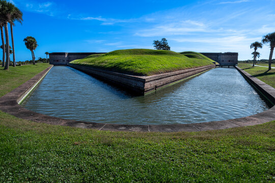The water-filled moat and grassy earthworks of Fort Pulaski lead toward its historic brick walls beneath a blue sky.