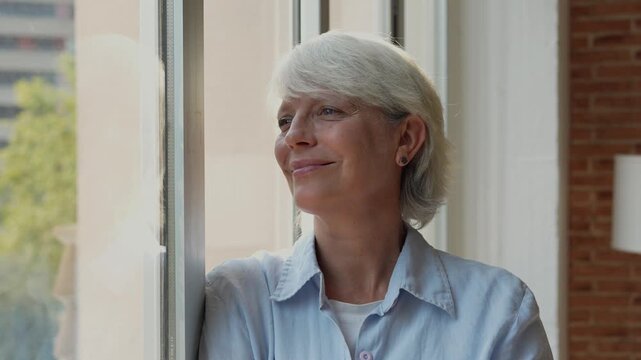 A beautiful senior woman with elegant grey hair enjoys a peaceful moment, smiling contently as she looks out a bright window. Her thoughtful expression radiates happiness and optimism for the future.