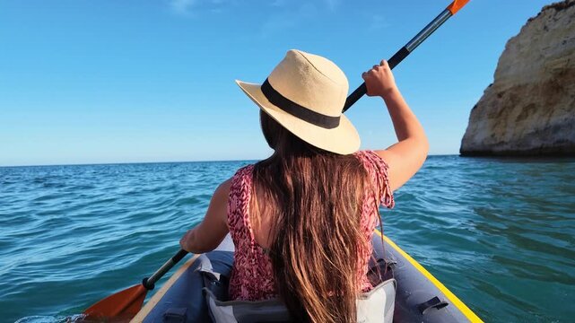 A young woman enjoys a tranquil kayaking adventure, paddling through the clear blue ocean waters under a sunny sky. She explores the stunning coastal landscape, passing by impressive rock formations.
