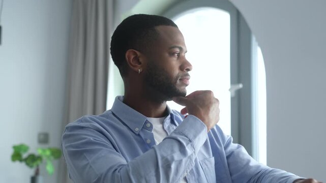 A thoughtful young African American man deeply contemplates, touching his chin as he processes ideas in a bright, modern indoor setting.
