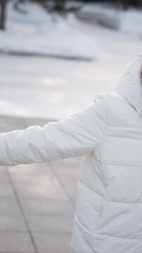 Woman wearing white puffer jacket black headphones and large sunglasses moves her arms while standing outdoors in winter. Her hands with painted nails are visible as woman gestures