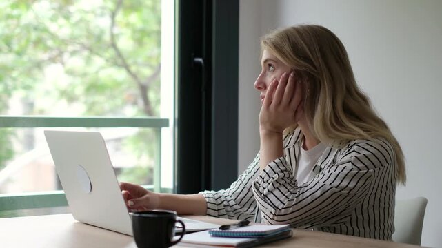 Young businesswoman feeling overwhelmed and tired while working on her laptop, displaying symptoms of burnout, stress, and lack of motivation in her modern home office by a window
