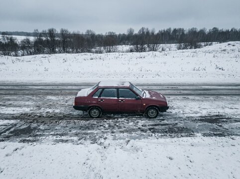 Old maroon sedan standing on the side of a dirty snowy road in winter landscape