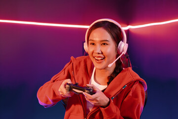 Host channel of smiling beautiful Asian girl streamer with joystick playing online game wearing headphones paste talking with viewers media online. Esport skilled team players in neon room. Stratagem. © InfiniteFlow