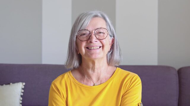 A cheerful senior lady with gray hair and glasses sits comfortably on a sofa, looking at the camera with a warm smile and a delightful laugh. Her happiness is evident.