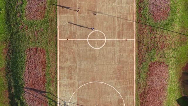 Concrete multi sport court features painted center circle and goal area, elongated shadows of players stretching across surface near Posadas, Misiones, drone flying forward top down.
