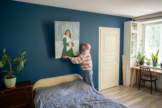 Creative woman hanging own painting on wall in bedroom at home carefully placing artwork, focused female standing by bed and attentively putting personal large drawn canvas art in apartment.