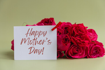 White greeting card with Happy Mother's Day message clipped in front of fresh red rose bouquet on soft green background. Floral composition with generous copy space suitable for Mother's Day greetings © anna.stasiia