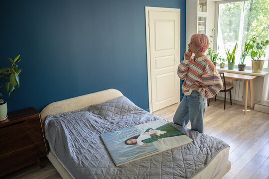 Thoughtful woman painter planning decorating wall with canvas painting in own bedroom, pensive female standing beside bed looking at wall space planning place for hanging masterpiece lying on bed.