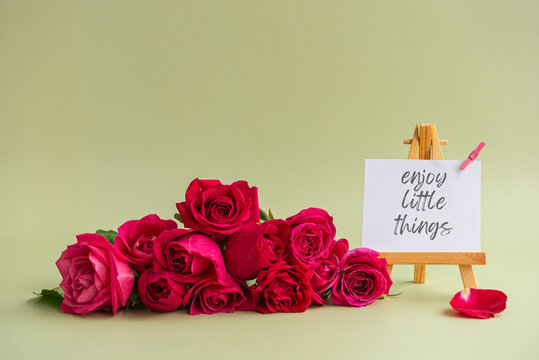 Inspirational quote card ENJOY LITTLE THINGS displayed on wooden easel surrounded by vibrant pink roses and loose petal against soft pastel background. Elegant floral composition with motivational