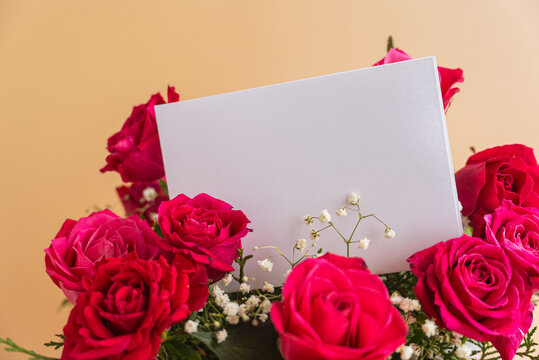 Pink gift box filled with fresh red and pink roses, greenery and delicate flowers holding blank greeting card envelope. Decorative ribbon and elegant floral arrangement create festive composition with