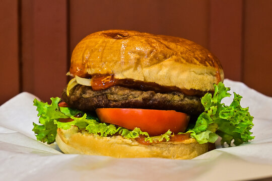 side view of burger with patty, sliced tomatoes, lettuce and other vegetables. front, close up. hamburger. home made. tasty.  beef, meat. fast food. gourmet. brown wall background. white paper.