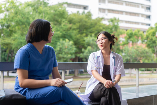 Young Asian female medical staff talking and relaxing outdoors at hospital, doctor and nurse in uniform having a break together outside hospital building