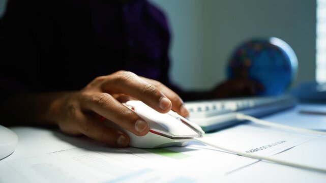 Professional working with documents at an office desk, Close-up of a professional hand using a computer mouse beside documents on a bright office desk, with a blue globe and window in the background