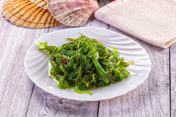 Fresh green seaweed salad on white plate with seashells and textured cloth on rustic wooden table © Andrei Starostin