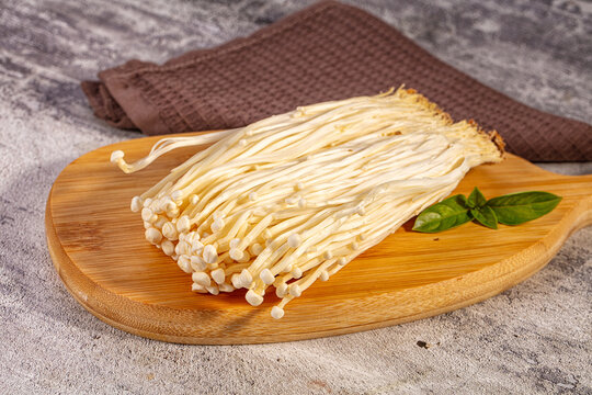 Fresh enoki mushrooms on wooden cutting board with basil leaves, light beige stems and small caps, rustic kitchen setting